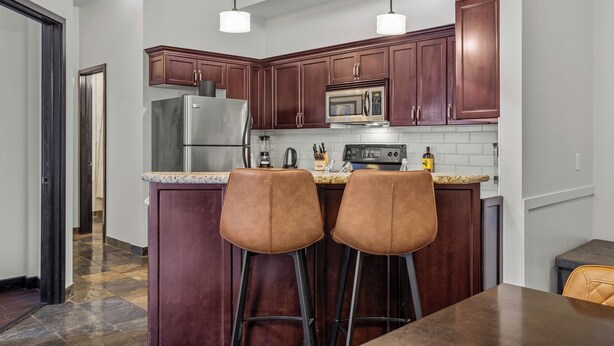 A modern kitchen with dark wooden cabinets, stainless steel appliances, and a granite countertop. There are two bar stools and pendant lights above the counter.