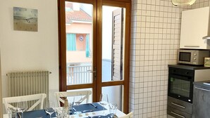 Un comedor con una mesa dispuesta para cuatro, sillas blancas, un reloj en la pared, vistas a través de un ventanal y una cocina con microondas y horno.