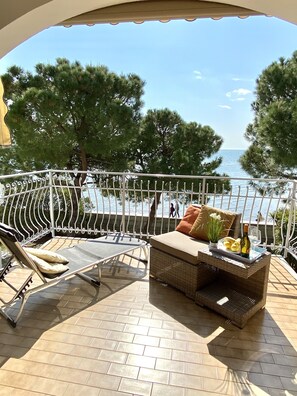 A balcony with a wicker sofa, a lounge chair, and a table with a bottle and glasses. - Seaview apartment at Costa Azzura beach (Grado)
