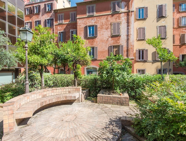 A courtyard with a brick archway, surrounded by greenery and buildings with multiple windows and shutters.