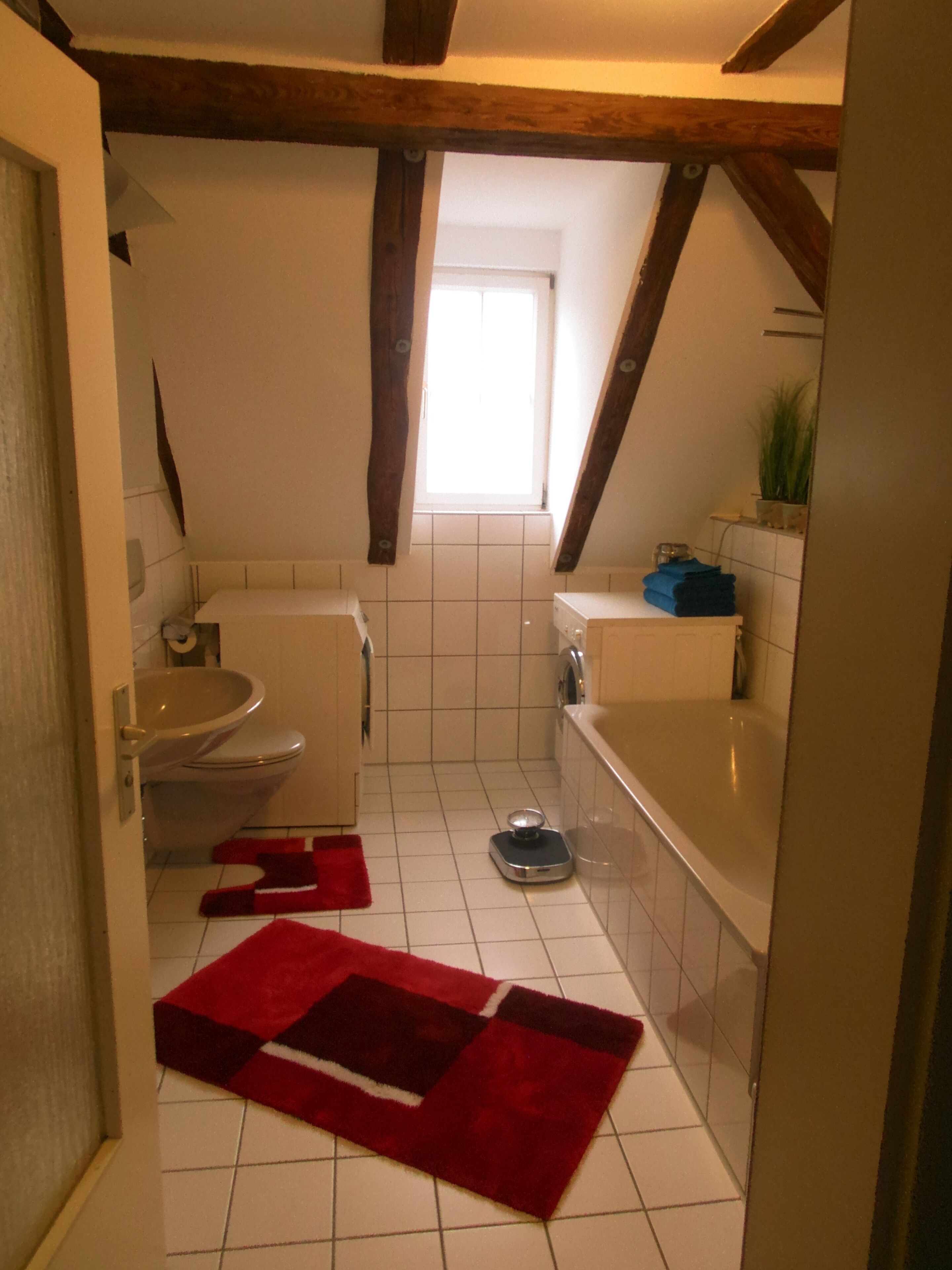 A small, narrow bathroom with a toilet, sink, and bathtub. There is a window providing natural light, and the floor is tiled.