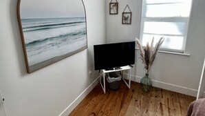 A room with a wooden floor, a large arched picture of the ocean, a flat-screen TV on a stand, a window with a view of the sea, and two small framed pictures on the wall. - Private Beach Front Getaway at The Sunshine Beach Cottage. (Scotland)