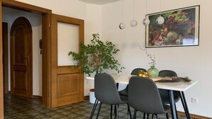 Ein Essbereich mit einem weißen Tisch, zwei schwarzen Stühlen, einer Topfpflanze und Wandbildern mit Obstmotiven.