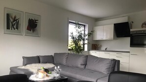 Ein modernes Wohnzimmer mit einem grauen Sofa, einem kleinen schwarzen Tisch mit Kerze und Weingläsern sowie einer Küchenzeile mit weißen Schränken und einem schwarzen Fernseher.