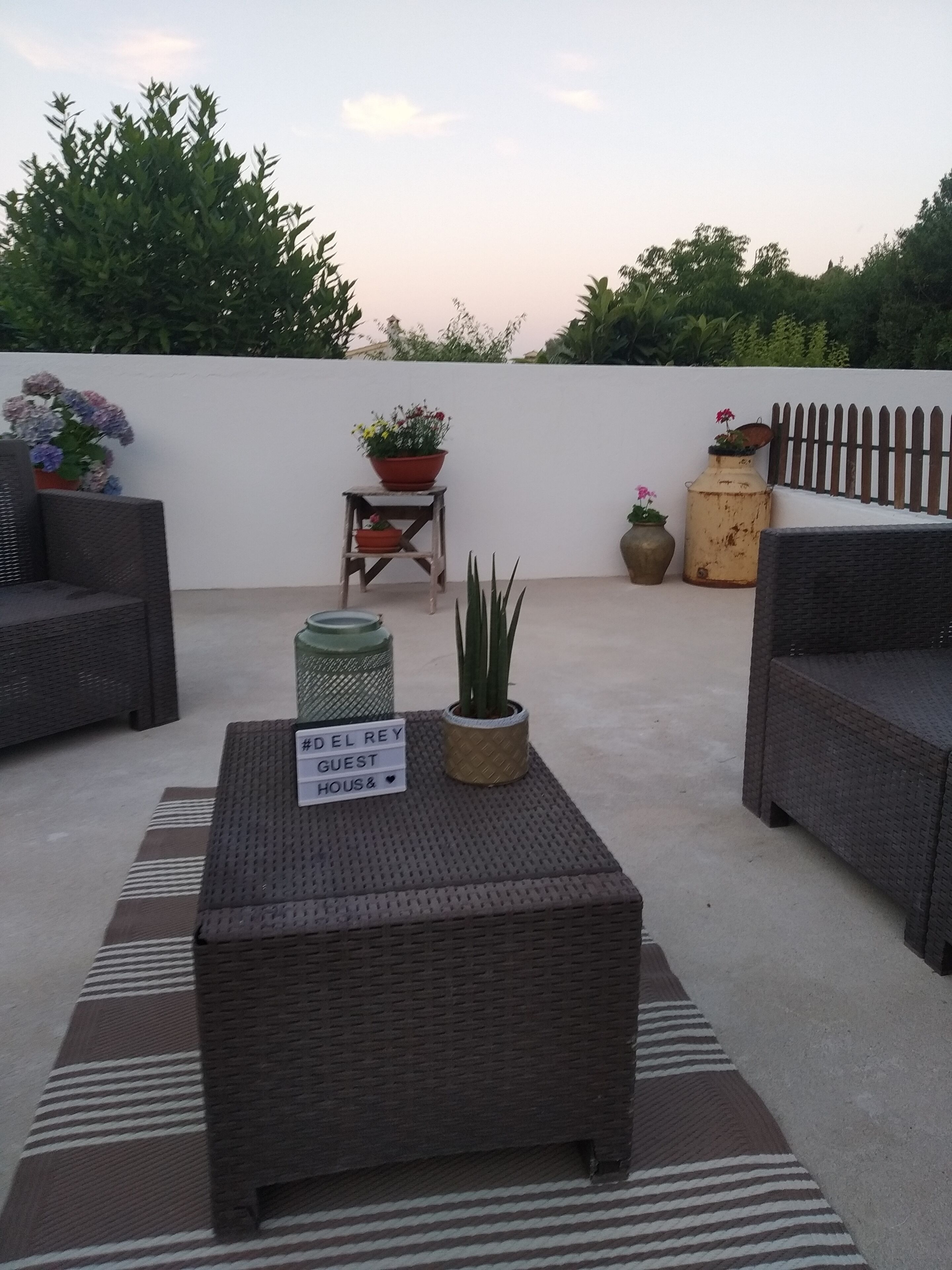 A rooftop patio with wicker furniture, a sign reading "#DEL REY GUEST HOUSE", and potted plants.