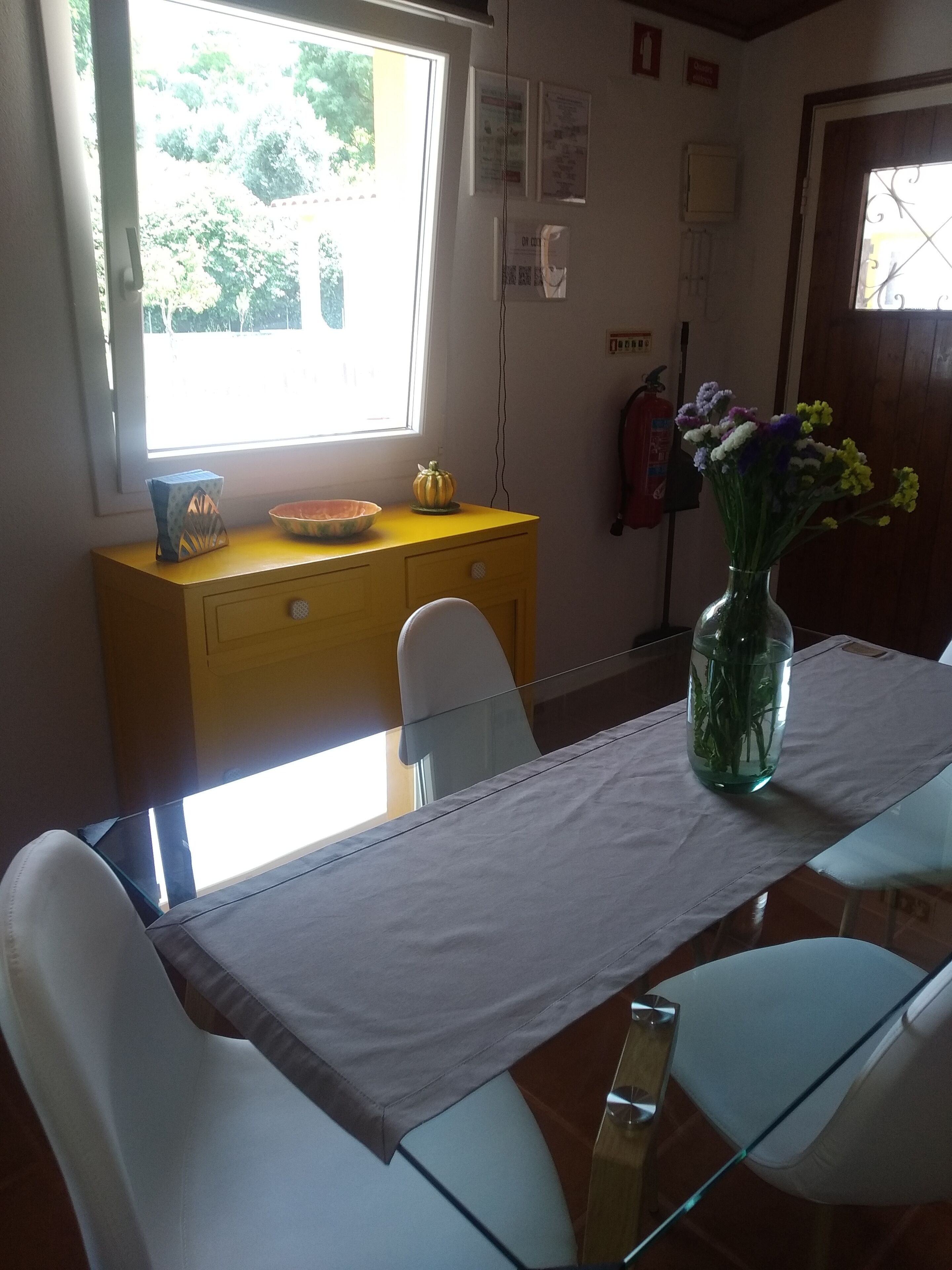 Un coin repas avec une table dressée pour deux personnes, un buffet jaune et un vase rempli de fleurs.