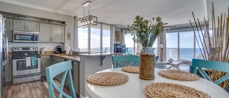 Une cuisine moderne dotée d’appareils électroménagers en acier inoxydable, un coin repas agrémenté de sets de table en fibres naturelles, et une vue sur l’océan.