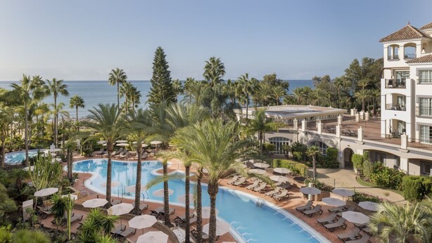 Un resort con piscina rodeada de palmeras y sillas de descanso, con vistas al océano.