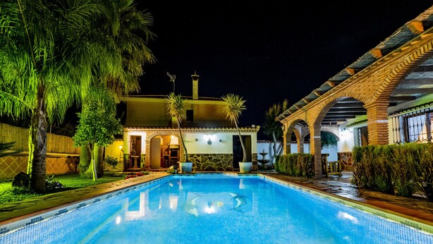 A swimming pool with a tiled deck, surrounded by palm trees and a building with a tiled roof and brick walls.