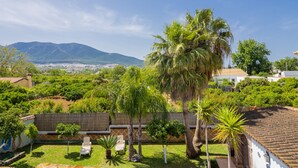 A residential area with a view of a mountain range, a fenced yard with palm trees, and a white building with a tiled roof.