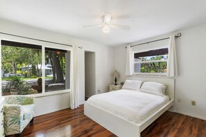 A bedroom with a bed, a nightstand, a lamp, a ceiling fan, and large windows with curtains.