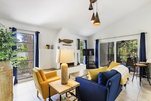 A spacious living area with a sofa, armchairs, a coffee table, and a TV. There is a large window with curtains, a plant, and a bookshelf.