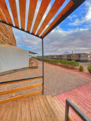 A covered outdoor area with a wooden deck, a pergola, and a view of a row of modern buildings in the background. - Rum Oasis Luxury Camp (Wadi Rum)