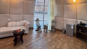 Een gezellige kamer met een comfortabele zithoek, twee kleine tafels en een koelkast. Een groot raam met gordijnen biedt uitzicht op de omgeving.