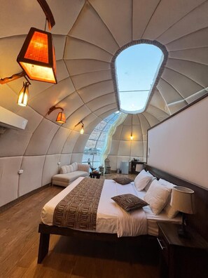 A modern, curved-roofed bedroom with a skylight, a large bed with patterned bedding, and ambient lighting fixtures. - Rum Oasis Luxury Camp (Wadi Rum)