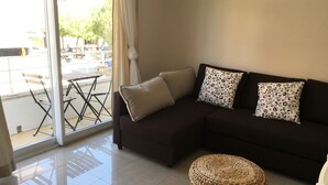 A living room with a dark brown sofa, two patterned pillows, a woven ottoman, and a glass door leading to an outdoor area with a table and chairs.