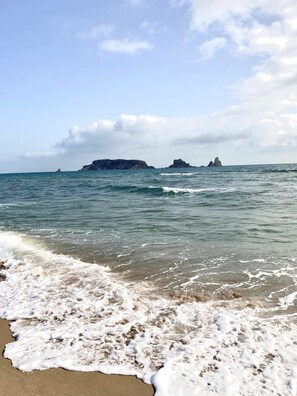 Une plage où les vagues déferlent sur le rivage, avec, au loin, des affleurements rocheux.