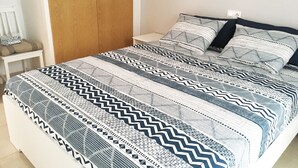 A neatly made bed with a patterned bedspread and pillows, a wooden wardrobe, and a white nightstand with a lamp.