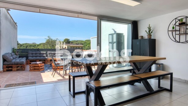 Un salon moderne doté d’une grande porte-fenêtre coulissante donnant sur une terrasse. L’intérieur comprend une table à manger en bois avec des bancs, un ensemble de canapés et un meuble contenant des objets décoratifs.