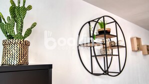 Un cactus dans un pot à motifs, posé sur un meuble noir, une étagère en métal noir avec une plante, et un mur blanc.