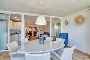 A dining area with a round glass table surrounded by wicker chairs, a hanging light fixture, and a view into the kitchen.