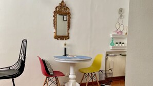 Un espace de vie compact comprenant un coin repas avec une table blanche et quatre chaises, un miroir décoratif, un radiateur et une petite étagère ornée d’objets décoratifs.