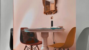 Un coin repas comprenant une table blanche sur pied, deux chaises, un miroir décoratif et une petite table avec une lampe.