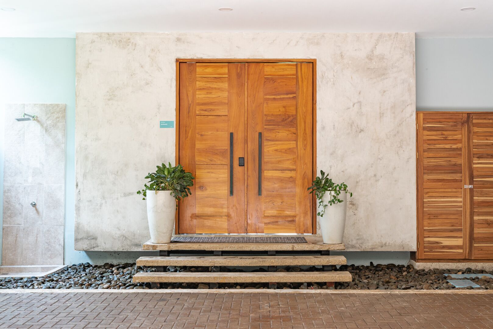 Una puerta doble de madera con un picaporte metálico, flanqueada por dos macetas, que da acceso a una escalera de piedra.