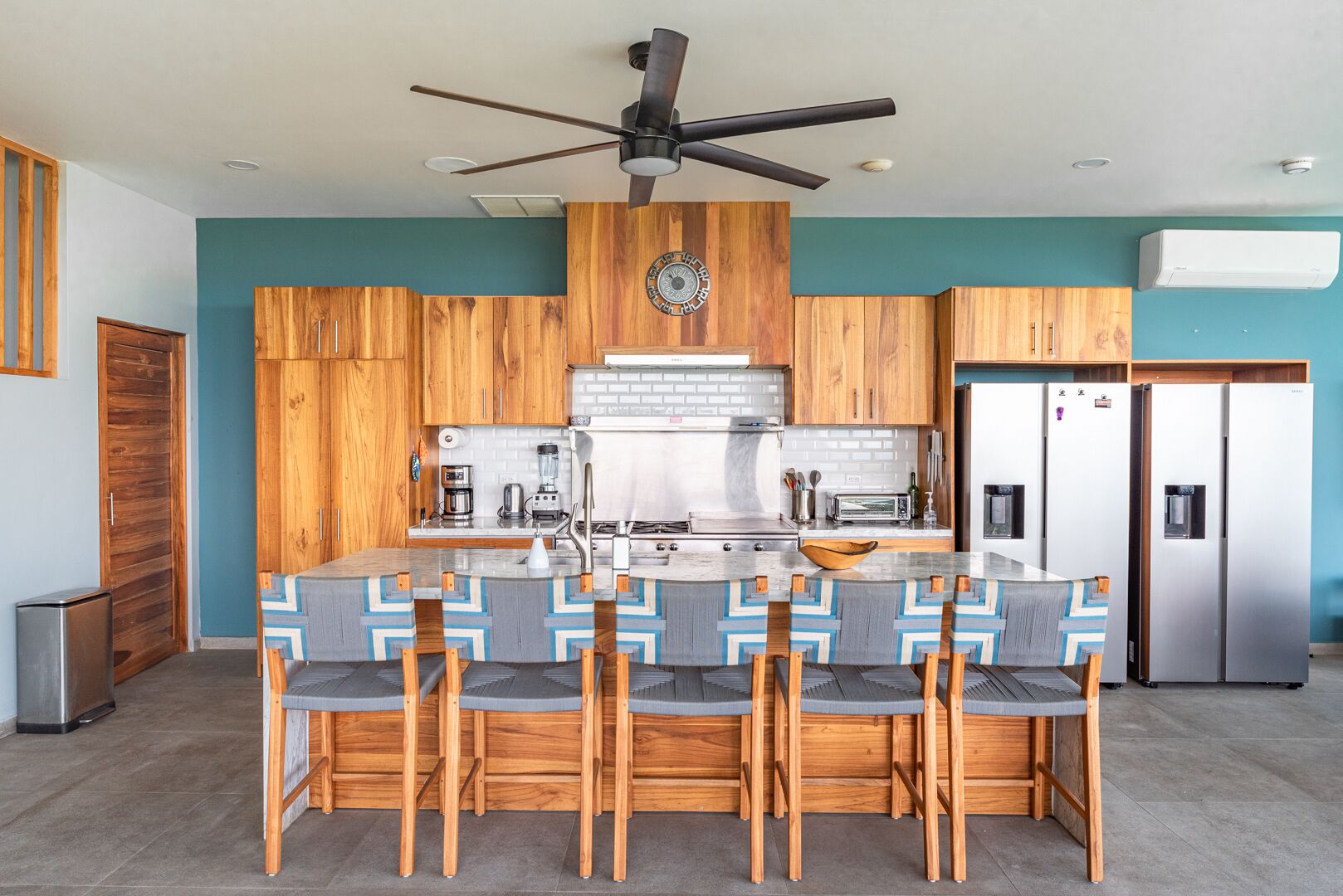 Una cocina moderna con armarios de madera, una isla central con taburetes de bar, ventilador de techo y electrodomésticos de acero inoxidable.
