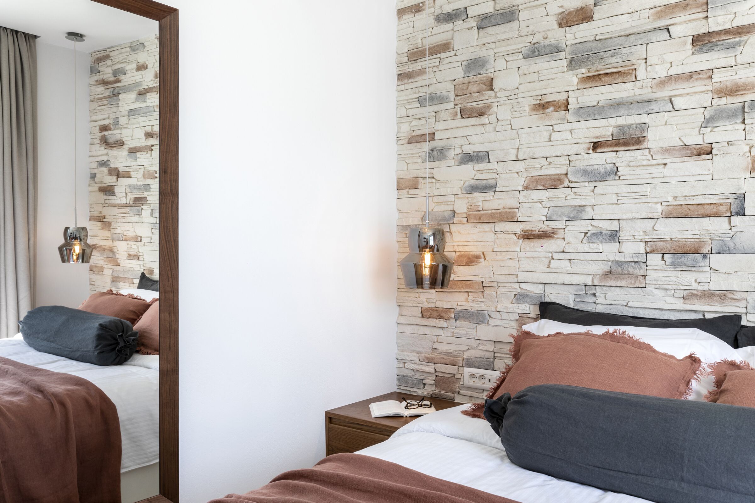 Una habitación de hotel con pared de piedra, una mesita de noche de madera con un libro y gafas, y una cama con ropa de cama marrón y gris.