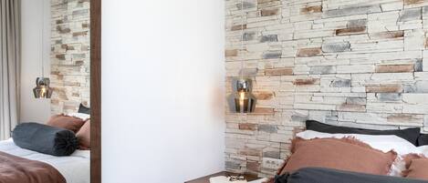 Una habitación de hotel con pared de piedra, una mesita de noche de madera con un libro y gafas, y una cama con ropa de cama marrón y gris.