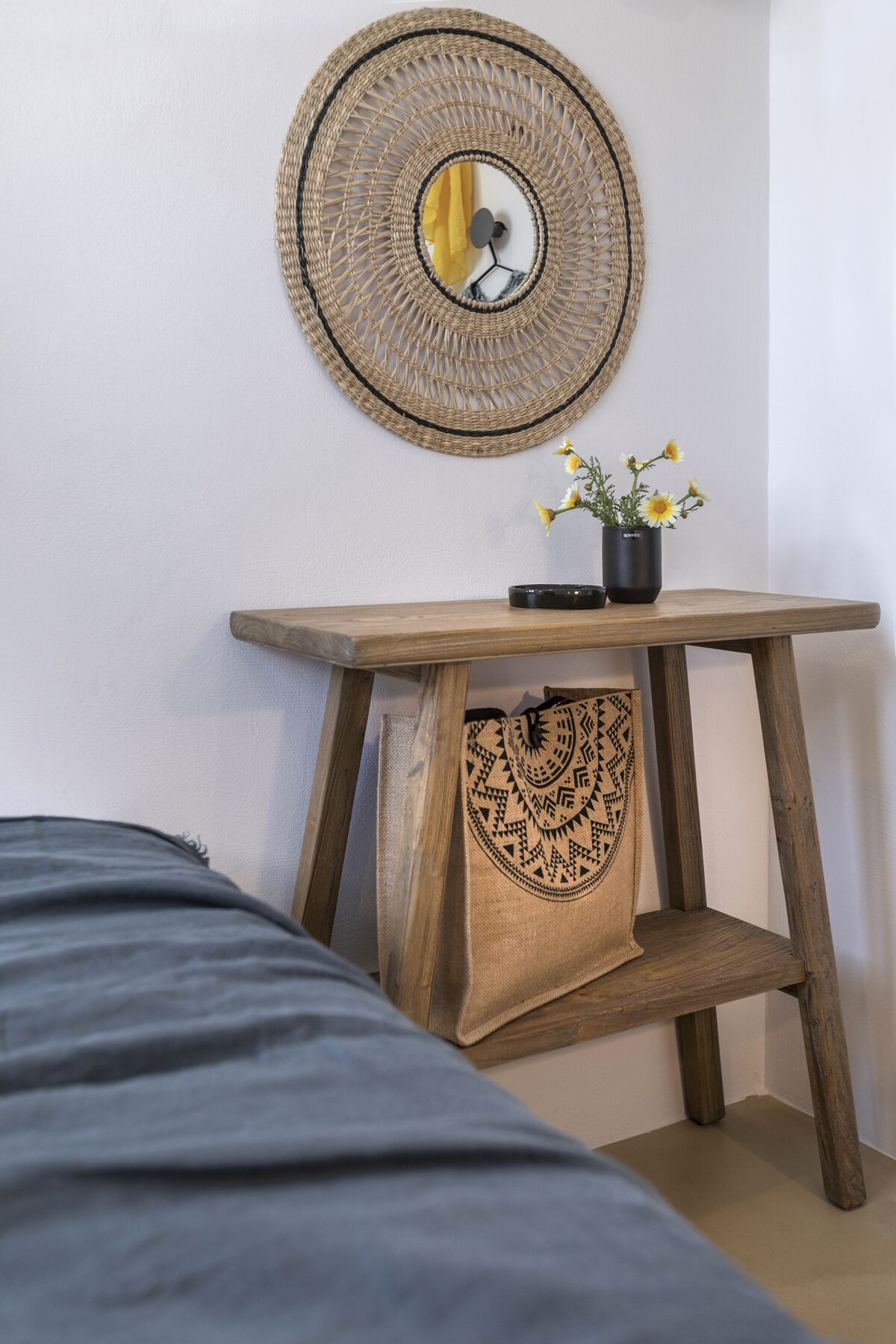 A room with a bed, a wooden bedside table with a woven bag, a mirror, and a vase with flowers.