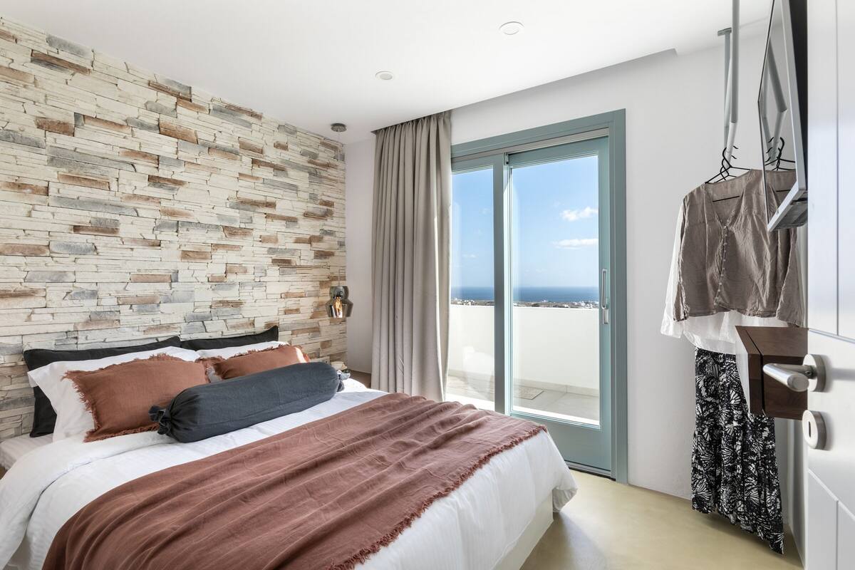 Quarto moderno com parede de pedra, uma cama grande com várias almofadas, vista para o mar através de uma porta deslizante e um cabideiro.