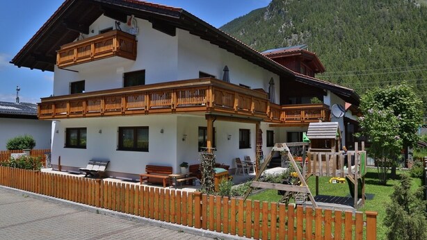 A two-story chalet with wooden balconies, a covered patio, and a play area for children. - Ferienwohnung in Traumhafter Bergkulisse - Appartements Wacker (Bichlbach)