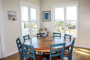 Une salle à manger avec une table ronde en bois, entourée de six chaises bleues, de grandes fenêtres et deux tableaux encadrés accrochés aux murs.
