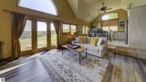 A spacious living area with a high ceiling, a white sofa, a coffee table, and a view of the outdoors through large windows. - Live the Vineyard Life at The Grand View 2 @ Willow Winery (Suttons Bay)