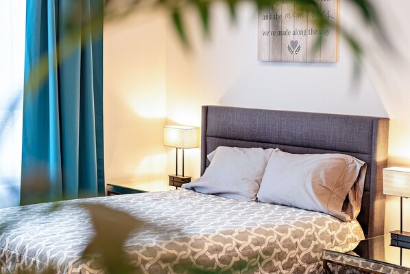 A neatly made bed with patterned bedding, a bedside lamp, and a wall-mounted sign with a heart and laurel wreath symbol. - #104 Unique Place Downtown Monroe! (Monroe)