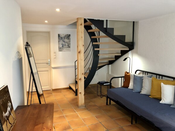 A room with a wooden floor, a bed with pillows, a small table, a mirror, and a staircase with a wooden handrail.