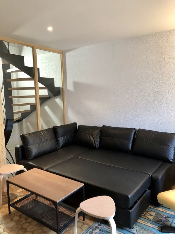 Un salón moderno con un sofá de cuero negro, una mesa de centro de madera y un taburete blanco. Se observa una escalera con escalones de madera y barandillas negras al fondo.