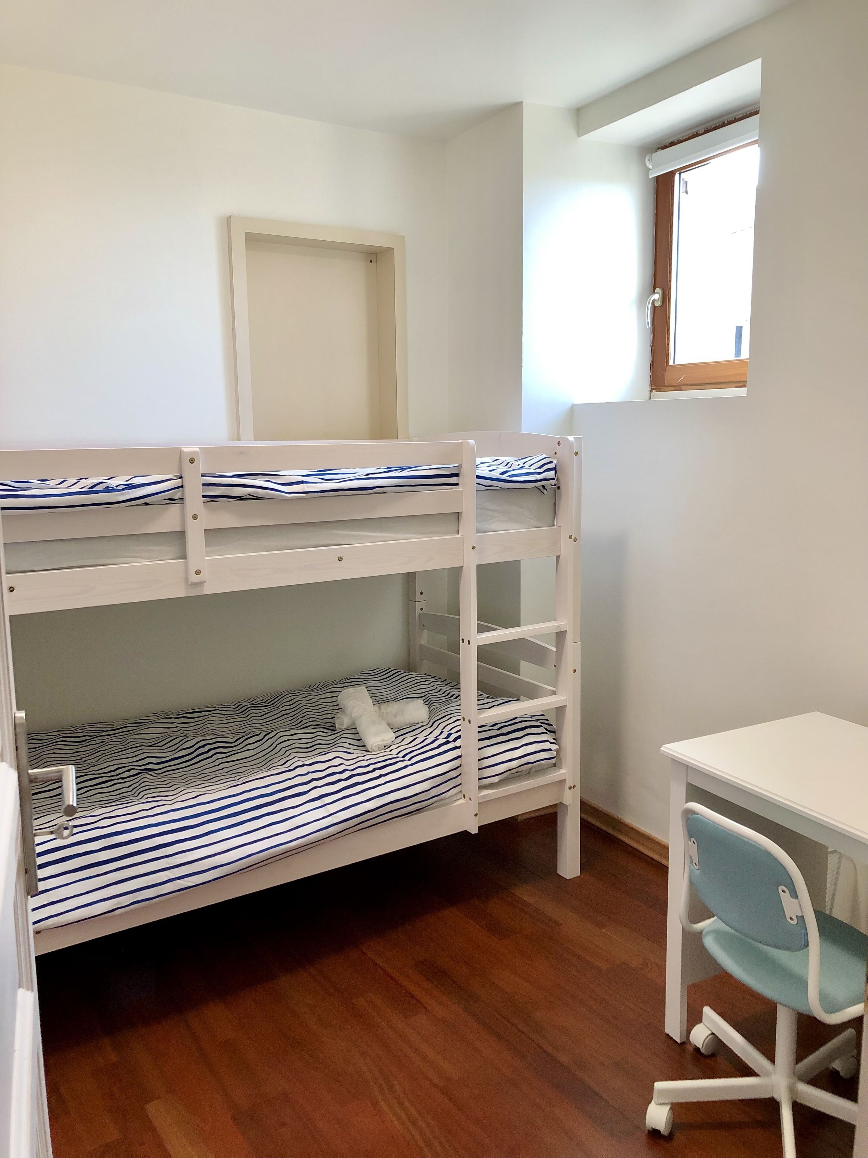 Un letto a castello con biancheria a righe, una scrivania bianca e una sedia in una stanza con pavimento in legno e una finestra.