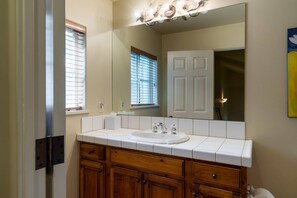 Un baño con lavamanos blanco, armarios de madera, un espejo grande y una ventana con persianas.