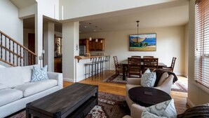 A spacious living area with a sofa, coffee table, and a dining table with chairs. A kitchen is visible in the background with wooden cabinets and a hanging light fixture. - Great Ketchum Condo next to bike path & greenbelt (Ketchum)