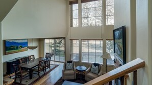 A spacious living area with a dining table, chairs, a sofa, and a coffee table. Large windows provide natural light and views of the outdoors. - Great Ketchum Condo next to bike path & greenbelt (Ketchum)