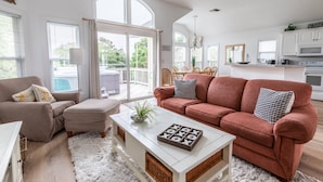A spacious living room with a large window, a red sofa, a white coffee table, and a kitchen area with white cabinets and appliances. - 1767 - Fox Snooze - Corolla Retreat | Pool, Hot Tub & Beach Walk (Corolla)