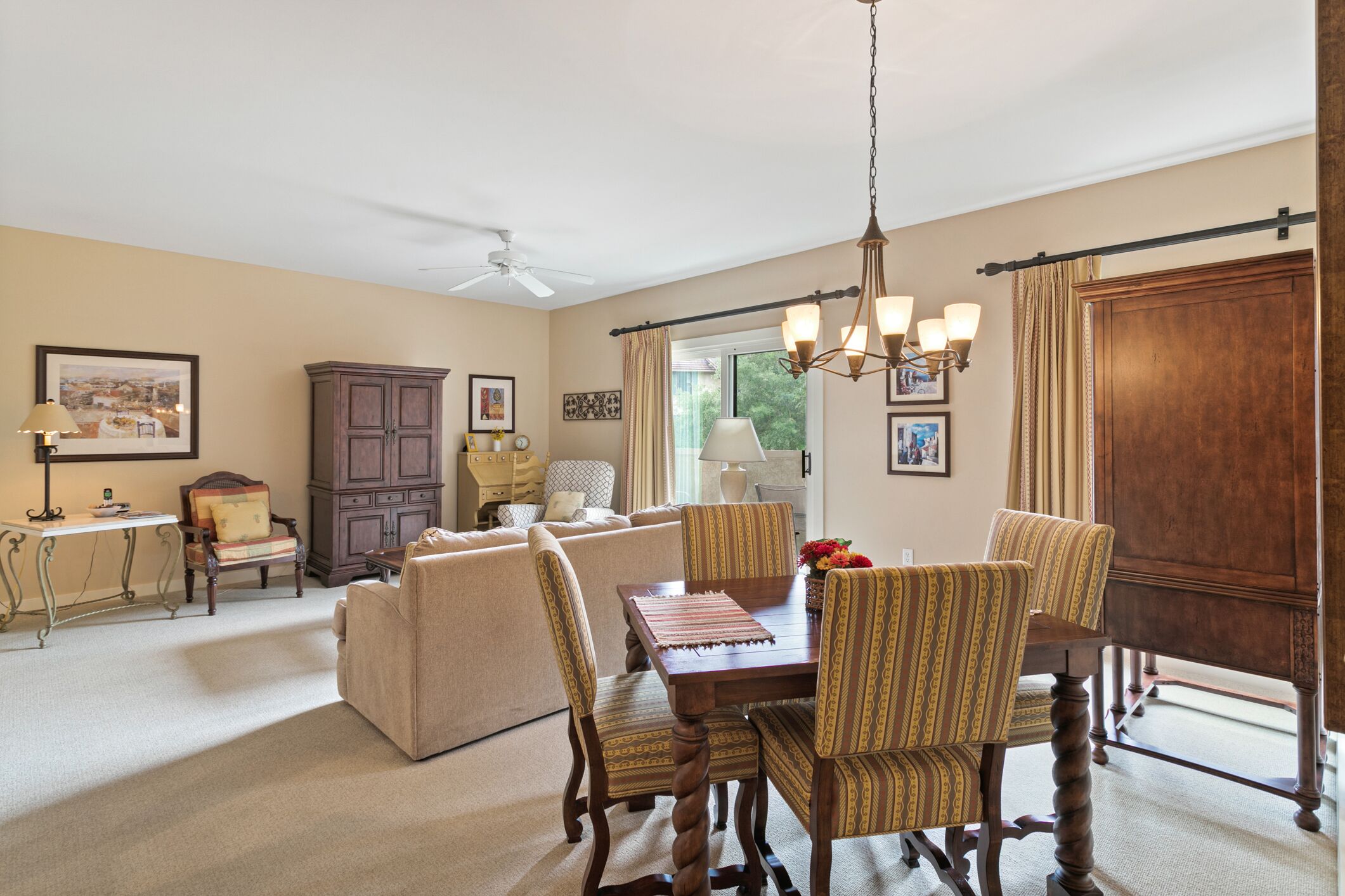 A dining area with a wooden table, patterned chairs, and a chandelier. There is a sofa and a cabinet in the background.