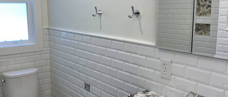 Une salle de bain avec des toilettes blanches, un lavabo doté d’un plan de travail en marbre et un miroir avec un pare-feu en forme de motifs de briques.