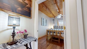 Een kamer met een houten plafond, een bed met een geruit beddegoed, een tafel met een lamp en bloemen, en een decoratie van een windmolen aan de muur.