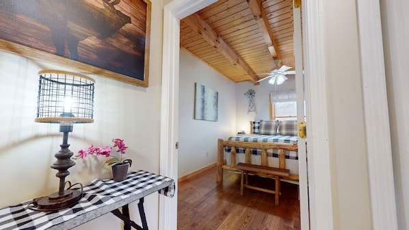 A room with a wooden ceiling, a bed with a checkered bedspread, a table with a lamp and flowers, and a windmill decoration on the wall.