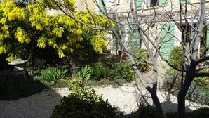 Ein Steinhäuschen mit grünen Fensterläden, umgeben von einem Garten mit blühenden gelben Bäumen und verschiedenen Sträuchern.