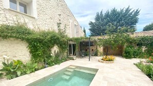 Ein Pool mit einer Steindeckplatte, umgeben von Grünpflanzen und einem Steinhaus.